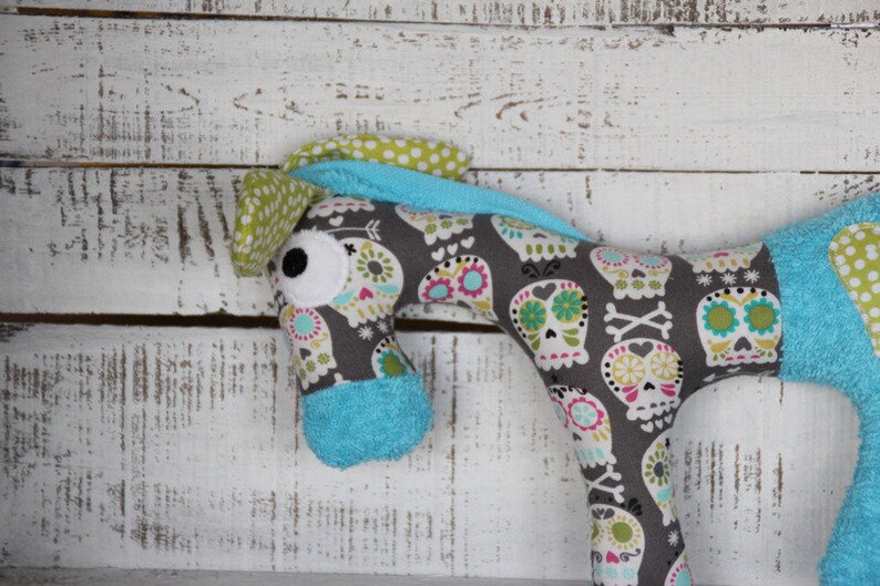 以下が含まれることがあります： 骸骨と花の模様がついたグレーとブルーのぬいぐるみ馬。馬は青と白のドットの鼻と大きな白い目を持っている。