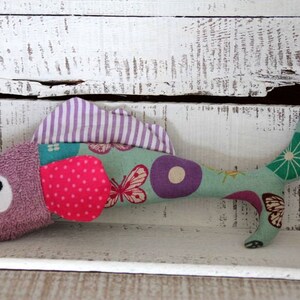 Könnte beinhalten: Ein lila-grünes, fischförmiges Stofftier mit einem rosa Tupfen-Patch und einem weißen Auge. Das Spielzeug hat einen gestreiften lila-weißen Stoff oben und einen grünen Stoff mit einem Schmetterlings- und Kreis-Muster unten. Das Spielzeug hat ein Etikett mit der Aufschrift "Lieb" darauf.