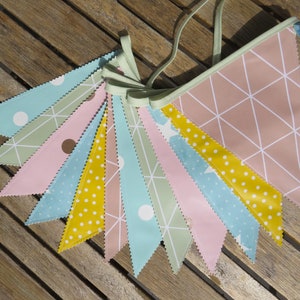 Może przedstawiać: Kolorowy girlanda z trójkątnymi flagami w pastelowych odcieniach różu, błękitu, żółci i zieleni. Flagi mają grochy, geometryczne wzory i wzory gwiazd. Girlanda jest związana jasną zieloną wstążką.