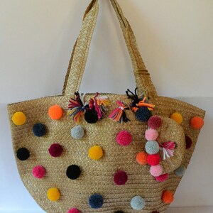 May include: A straw tote bag with colorful pom-poms in various colors, including yellow, orange, pink, and blue. The bag has a matching small pouch attached and gold-colored handles.