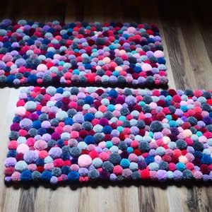 May include: Two rectangular rugs made of colorful pom-poms. The rugs are made of various shades of pink, purple, blue, and gray pom-poms.