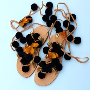 May include: A pair of tan leather sandals with black pom-poms, gold sequins, and orange and black floral accents. The sandals have long, tan leather straps for wrapping around the ankles. The sandals are displayed on a white surface.