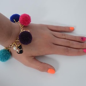 May include: A gold chain bracelet with colorful pom-pom charms. The bracelet has a purple, pink, blue, and teal pom-pom charm. The bracelet is on a person's wrist.