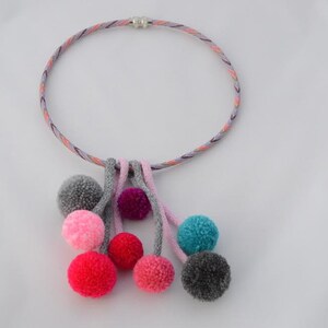 Puede incluir: Un collar con un cordón trenzado y un cierre plateado, con un grupo de pompones coloridos colgando de la parte inferior.