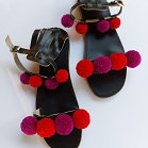 May include: Black leather sandals with multiple red and purple pom-poms attached to the straps.