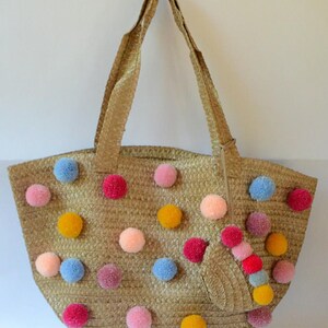 May include: A straw tote bag with a zipper closure and pom poms in various colors, including pink, blue, yellow, and red.
