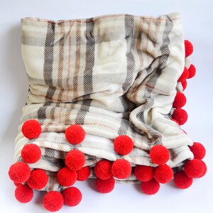 Peut inclure: Une couverture en plaid blanc et marron avec une bordure de pompons rouges. La couverture est pliée et les pompons sont visibles sur le bord inférieur.