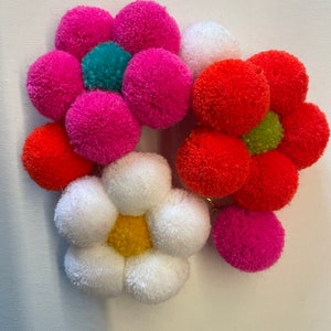 May include: A set of five colorful pom-pom flowers. The flowers are made of yarn and have a variety of colors, including pink, red, white, and green. The flowers are arranged in a cluster.