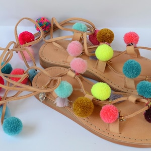 May include: A pair of tan leather sandals with colorful pom-poms attached to the straps. The sandals have a lace-up design and are tied around the ankles.
