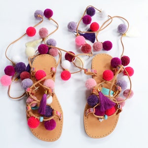 May include: A pair of brown leather sandals with multi-colored pom-poms and tassels. The sandals have a criss-cross strap design and are tied up the legs with strings.