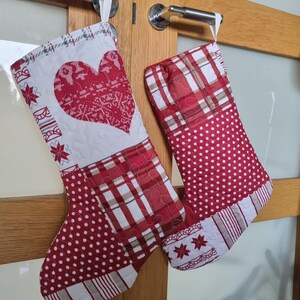 Può includere: Due calze di Natale appese a una porta di legno. Una calza presenta un grande cuore rosso con un motivo a fiocco di neve bianco. L'altra calza ha un motivo a quadri e pois rossi e bianchi. Entrambe hanno passanti bianchi.