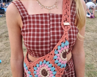 Op de afbeelding: Een vrouw draagt een rood-wit geruit jurkje met een gehaakte tas over haar schouder. De tas is gemaakt van bruin, roze, blauw en beige garen en heeft een groot cirkelvormig ontwerp.