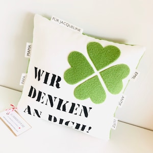 Könnte beinhalten: Quadratisches weißes Kissen mit einem großen Kleeblatt-Design aus grünem Filz. Das Kissen trägt den schwarzen Text "WIR DENKEN AN DICH!". Kleine Etiketten mit Namen sind an den Seiten angenäht.