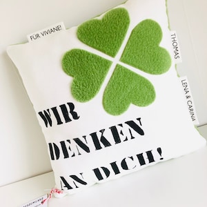Könnte beinhalten: Ein weißes Kissen mit einem grünen vierblättrigen Kleeblatt-Design und dem Text "WIR DENKEN AN DICH!" in Schwarz. Das Kissen hat ein Etikett mit der Aufschrift "FÜR VIVIANE!" und ein weiteres Etikett mit der Aufschrift "THOMAS LENA & CARINA".