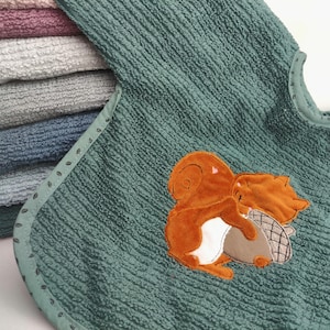 Könnte beinhalten: Ein türkisfarbenes Baby-Lätzchen mit strukturierter Oberfläche und einem braunen Eichhörnchen-Applikation, das eine Nuss hält. Das Lätzchen hat eine hellgrüne Paspel mit kleinen, dekorativen Nähten. Im Hintergrund ist ein Stapel gefalteter Handtücher in verschiedenen Pastellfarben zu sehen.