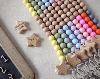 Puede incluir: Una colección de llaveros con filas de cuentas de silicona de colores y cuentas de madera natural. Cada llavero termina con una estrella de madera. Dos estrellas de madera sueltas están grabadas con el texto Schulkind 2026 y Sophie.