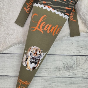 Könnte beinhalten: Olivgrüne Schultüte mit Tiger-Illustration und dem Namen "Leano" in Orange. Die Tüte hat eine passende Kapuze und Ärmel mit Zebrastreifenmuster. Der Text "Schulkini 2020" ist ebenfalls sichtbar.