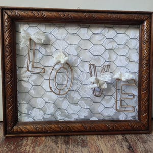 May include: A framed piece of art featuring the word "LOVE" spelled out in wire and decorated with white lace and flowers. The background is a piece of chicken wire.