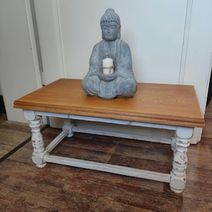 May include: A whitewashed wooden coffee table with a distressed finish and turned legs. A gray stone Buddha statue sits on the table top, holding a small white candle.