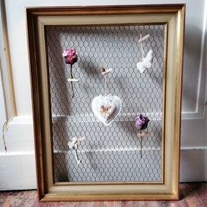 May include: A decorative framed display with a gold-colored frame and chicken wire backing. The display holds several small decorative items, including fabric hearts, roses, and keys, all attached with small clothespins.