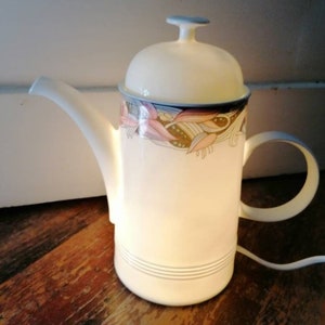 Könnte beinhalten: Eine weiße Teekanne aus Keramik mit einem Blumendesign und einem Licht im Inneren. Die Teekanne hat einen Deckel und einen Griff.