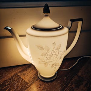 Könnte beinhalten: Eine weiße Porzellan-Teekanne mit floralem Muster, von innen beleuchtet, wodurch ein warmes Leuchten entsteht. Die Teekanne hat einen silberfarbenen Knopf auf dem Deckel und ein Kabel zum Anschließen.