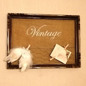 May include: A brown wooden frame with a burlap background. The word "Vintage" is written in white paint on the burlap. There are two white feathers with a small white heart attached to one of the feathers. A piece of paper with writing on it is pinned to the burlap with a small key.