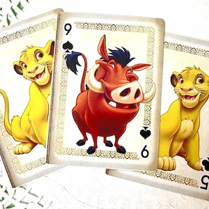 May include: Three playing cards featuring cartoon characters. The left card shows a yellow lion cub with the number 5 and a spade suit. The center card shows a red warthog with the number 9 and a spade suit. The right card shows a lion cub with a 5.