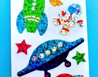 May include: A sheet of space-themed stickers featuring a green rocket, a silver astronaut, a blue flying saucer, a red planet with rings, a yellow crescent moon, a yellow comet, and three stars in red and green, all with a holographic glitter finish.