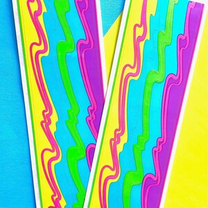 May include: Two colorful bookmarks with a repeating abstract design. The design features vertical bands of yellow, pink, green, blue, and purple. The bookmarks are on a blue and yellow background.