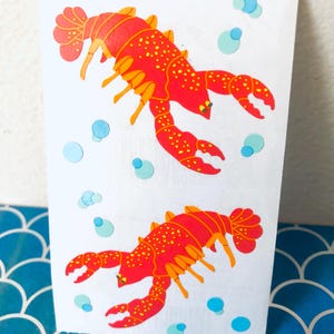 May include: White paper bag featuring two red and orange lobster illustrations with yellow spots and blue bubbles. The lobsters are in a swimming pose. The bag is on a blue and white patterned surface.