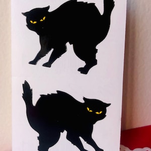May include: Two black cat stickers with yellow eyes on a white background. The cats are in a defensive posture, with arched backs and raised tails. The stickers are likely for Halloween or a cat-themed product.