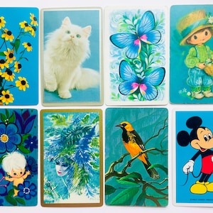 May include: A collection of vintage playing cards featuring various illustrations. The cards showcase images of flowers, a white cat, butterflies, a child with a basket, a bird, a cartoon character, and a woman with a blue headdress, all on a blue background.