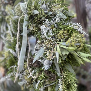 Könnte beinhalten: Ein handgefertigter Kranz aus verschiedenen grünen und silberfarbenen Blättern, darunter Eukalyptusblätter, Tannenzapfen und Beeren. Ein hellgrünes Band und Bindfaden werden zum Aufhängen verwendet. Eine natürliche, rustikale Dekoration.