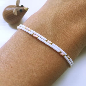 Könnte beinhalten: Zwei zarte, weiße Perlenarmbänder mit goldenen Akzenten und kleinen rosa Perlen werden an einem Handgelenk getragen. Im Hintergrund befindet sich eine kleine Holzmausfigur. Die Armbänder sind ein minimalistisches Schmuckdesign.