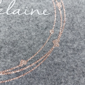 以下が含まれることがあります： グレーのフェルトポーチに白い文字で「Elaine」と書いてあります。ポーチには、ローズゴールドのグリッターでできた3つの同心円と、小さなローズゴールドのハートがアクセントになっています。