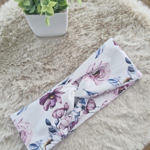 Könnte beinhalten: Ein weißes Stirnband mit einem Blumenmuster in Rosa-, Lila- und Blautönen. Das Stirnband ist verdreht und hat einen Knoten in der Mitte.