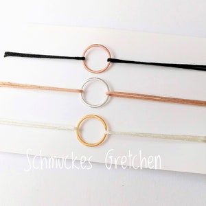 Könnte beinhalten: Drei Armbänder mit dünnen, kreisförmigen Metallringen in Roségold, Silber und Gold, jeweils an einer schwarzen, beigen und weißen Kordel befestigt. Der Text Schmuckes Gretchen ist unten auf der weißen Präsentationskarte aufgedruckt.