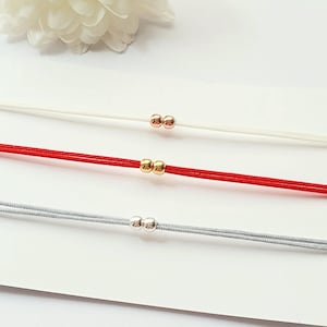 Könnte beinhalten: Drei Kordelarmbänder, die horizontal angeordnet sind. Das obere Armband ist weiß mit zwei roségoldenen Perlen, das mittlere ist rot mit zwei goldenen Perlen und das untere ist hellgrau mit zwei silbernen Perlen. Jedes Armband hat eine dünne Kordel.