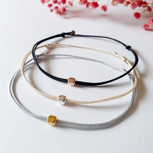 Könnte beinhalten: Drei Freundschaftsarmbänder mit verschiedenen Schnüren und quadratischen Perlen. Das obere Armband ist schwarz mit einer roségoldenen Perle, das mittlere Armband ist cremefarben mit einer silbernen Perle und das untere Armband ist grau mit einer goldenen Perle.