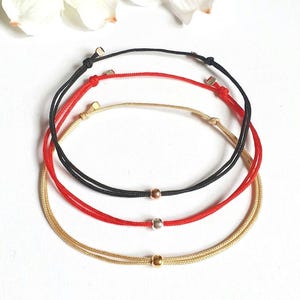 Könnte beinhalten: Drei verstellbare Kordelarmbänder in Schwarz, Rot und Gold. Jedes Armband verfügt über eine einzelne Metallperle in der Mitte und zwei kleinere Metallperlen in der Nähe des Schiebeknotenverschlusses, gefertigt aus dünnem, strapazierfähigem Material.