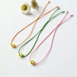 Könnte beinhalten: Drei verstellbare Kordelarmbänder in Orange, Grün und Pink, die jeweils eine einzelne gelbe Perle mit einem schwarzen Smiley-Gesicht aufweisen. Die Armbänder enthalten kleine Metallperlen an den Kordeln und sind neben zwei weißen Gänseblümchen abgebildet.