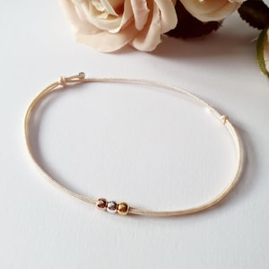 Könnte beinhalten: Ein zartes Armband mit einer dünnen, hellbeigen Kordel und einem verstellbaren Schiebeknotenverschluss. In der Mitte der Kordel befinden sich drei kleine, glatte, kugelförmige Perlen in den metallischen Ausführungen Roségold, Silber und Gelbgold.