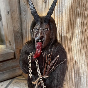 May include: A dark brown, furry figure with horns, a long red tongue, and a chain. The figure is holding a bundle of sticks and is standing on a wooden base.