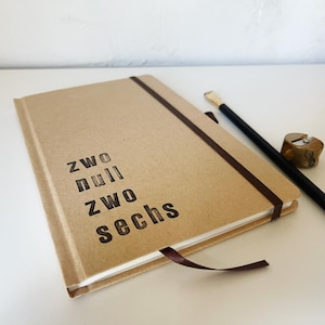 May include: A tan notebook with the text "zwo null zwo sechs" printed on the cover. A black pencil and a wooden pencil sharpener are next to the notebook. The notebook has a brown elastic band and a ribbon bookmark.