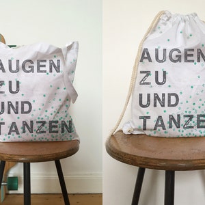 Puede incluir: Dos bolsas de lona blancas con texto negro y puntos verdes. El texto en las bolsas dice "Augen zu und Tanzen".