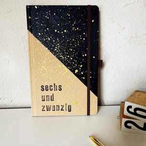 Könnte beinhalten: Ein Notizbuch mit einem schwarzen und goldenen, gesprenkelten Design auf der oberen Hälfte und einer beigefarbenen unteren Hälfte. Der Text "sechs und zwanzig" ist auf dem beigefarbenen Abschnitt aufgedruckt. Ein braunes Gummiband sichert das Notizbuch.