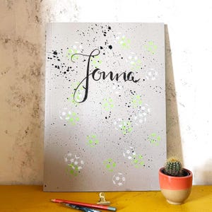 Puede incluir: Una libreta gris con el nombre "Jonna" escrito en caligrafía negra. La libreta está decorada con salpicaduras de pintura negra y diseños de balones de fútbol blancos y verdes.