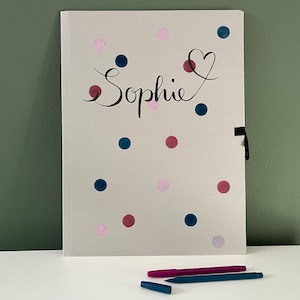 Könnte beinhalten: Ein weißer Ordner mit einem schwarzen Bandverschluss. Der Ordner hat ein Punkte-Muster mit rosa, blauen und burgunderfarbenen Punkten. Der Name "Sophie" ist in schwarzer Kursivschrift auf der Vorderseite des Ordners geschrieben.