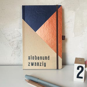 May include: A hardback notebook featuring a geometric design in navy blue, copper, and tan. The cover displays the text "siebenund zwanzig" in black. A dark brown elastic band secures the notebook, which includes a matching pen loop on the side.